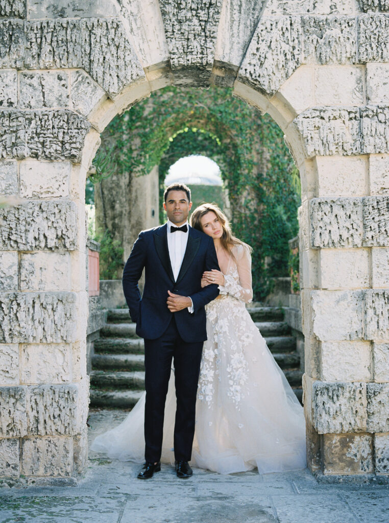 Bride resting her head on the grooms shoulder under a stone arch at Vizcaya.