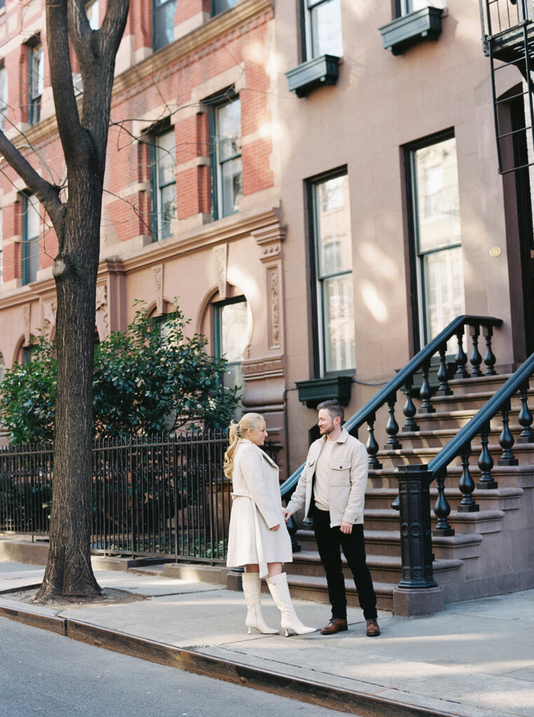 The couple is just engaged and standing in front of a brown stone on the west side in nyc.