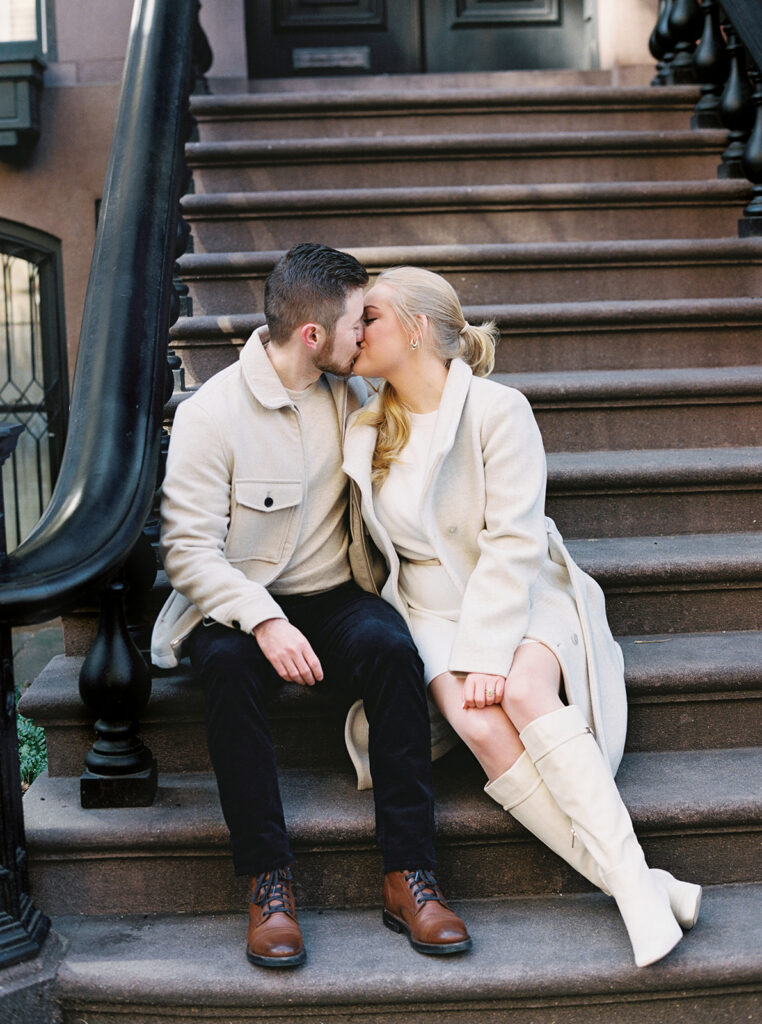 The couple is sitting on the brownstone stairs kissing.