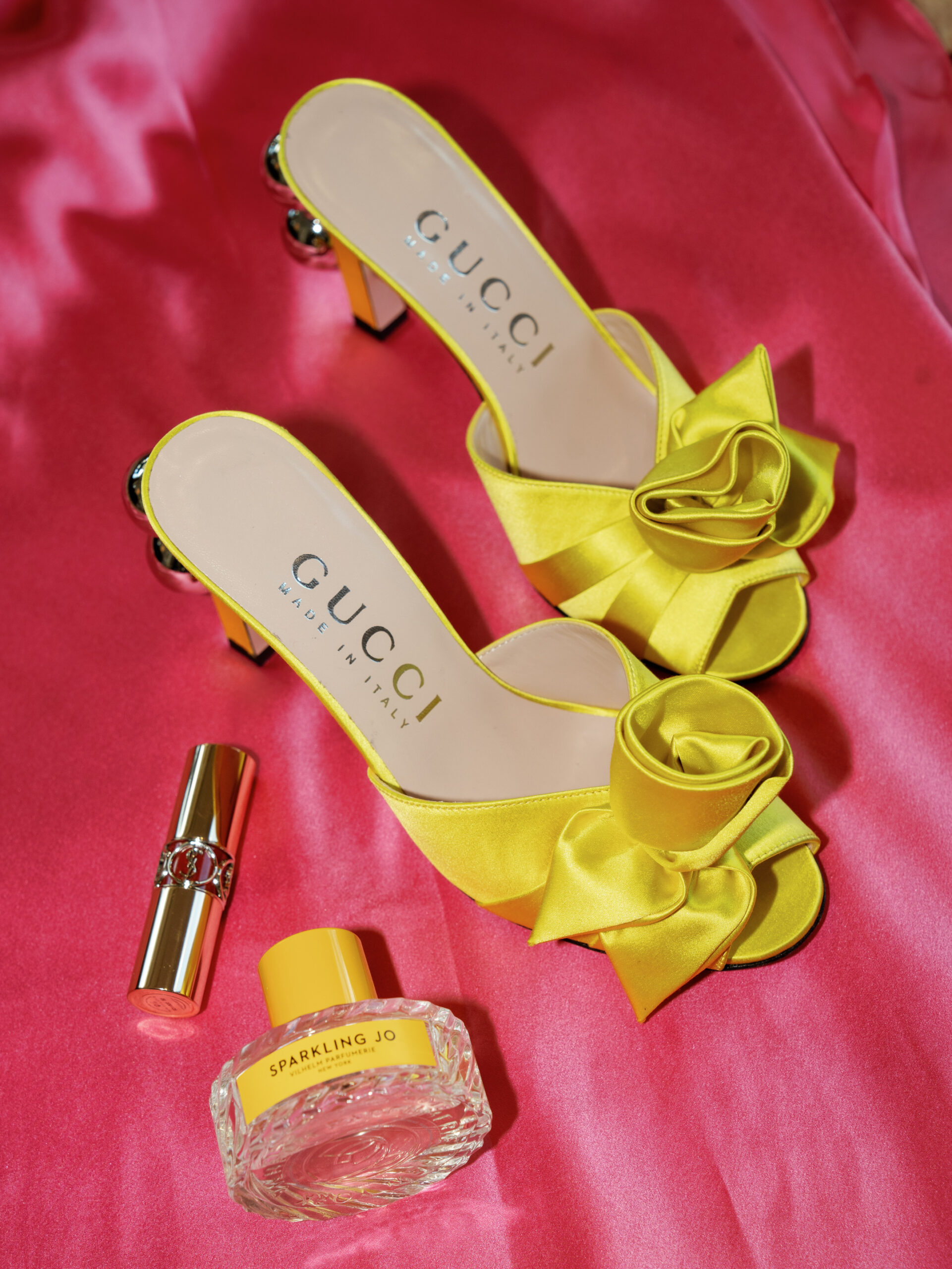 detail shot of the shoes, perfume and lipstick against a pink background