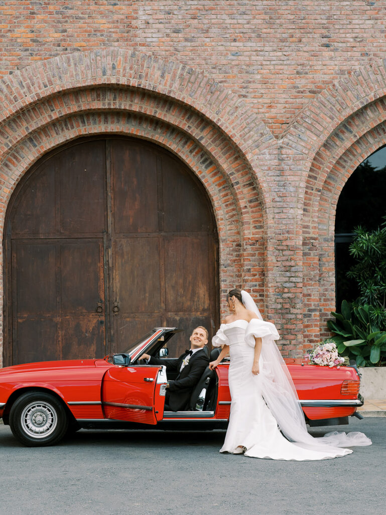 Bride and groom taking a portrait in front of red convertible.