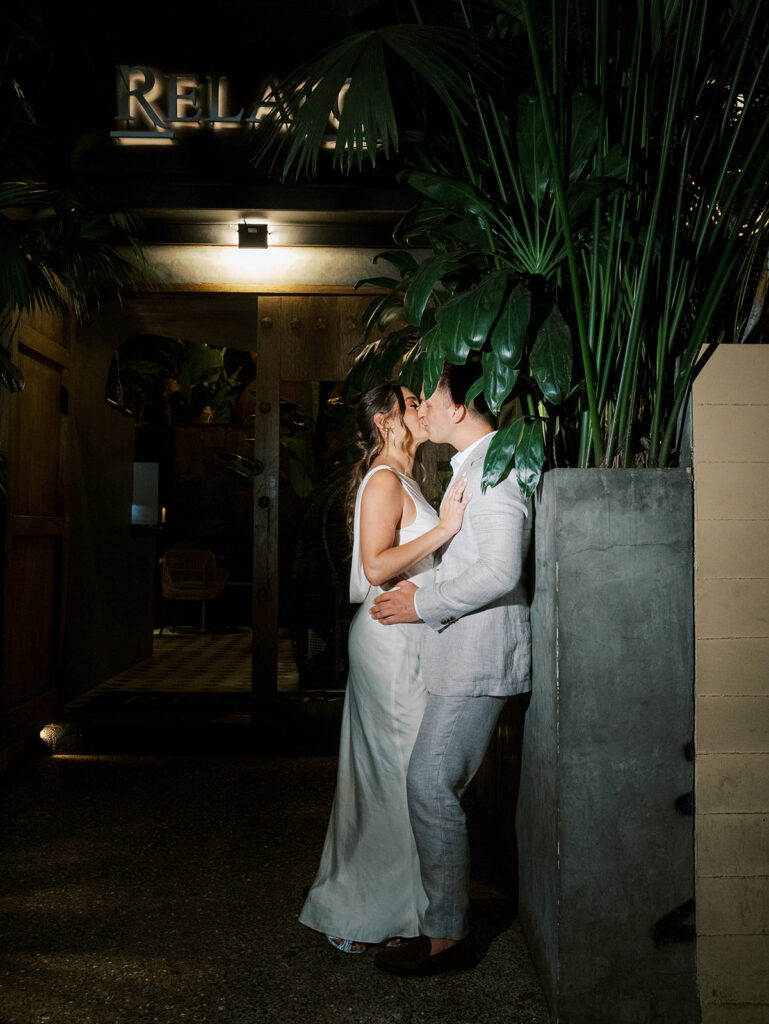 The bride and groom up against a wall outside of the restaurant kissing.
