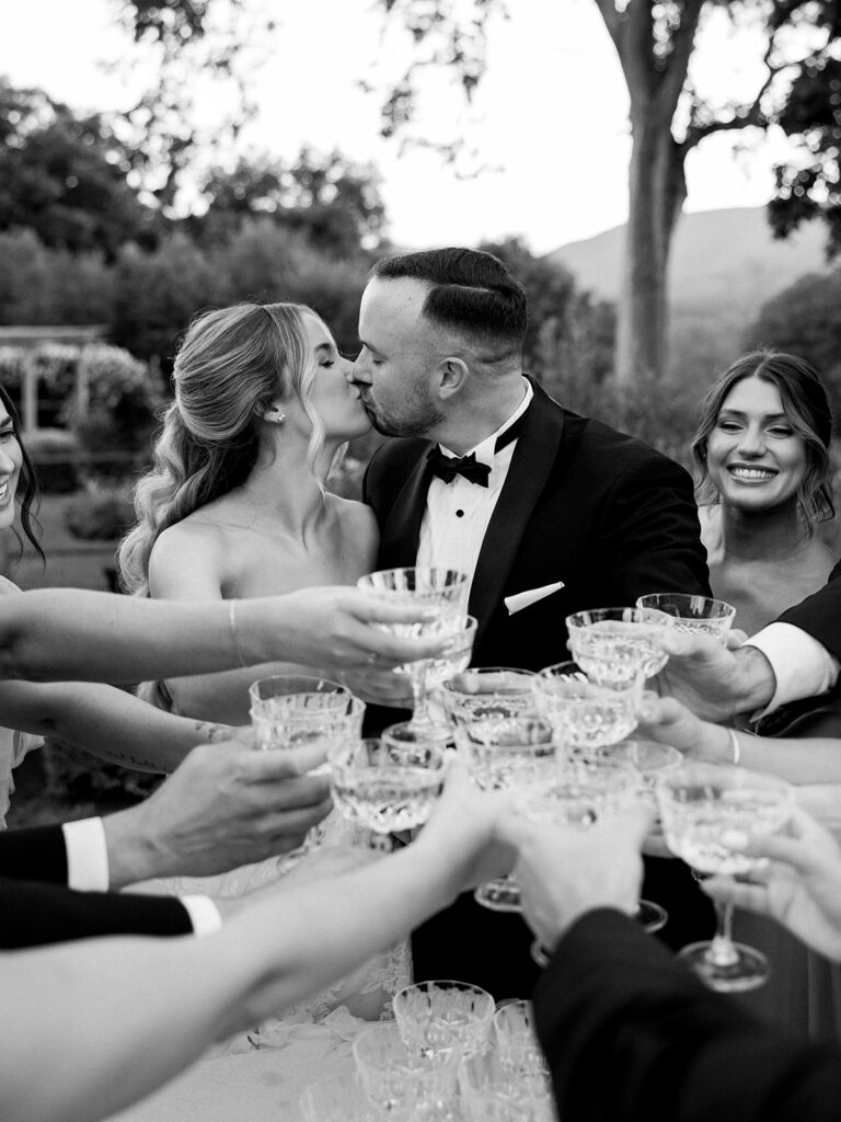 The bride and groom kissing during their champagne toast surrounded by the wedding party.