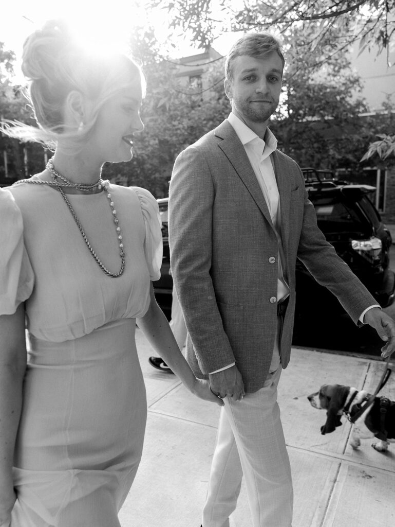 The bride and groom walking hand in hand with the groom looking at the camera. They are backlit with the sun peeking between them.