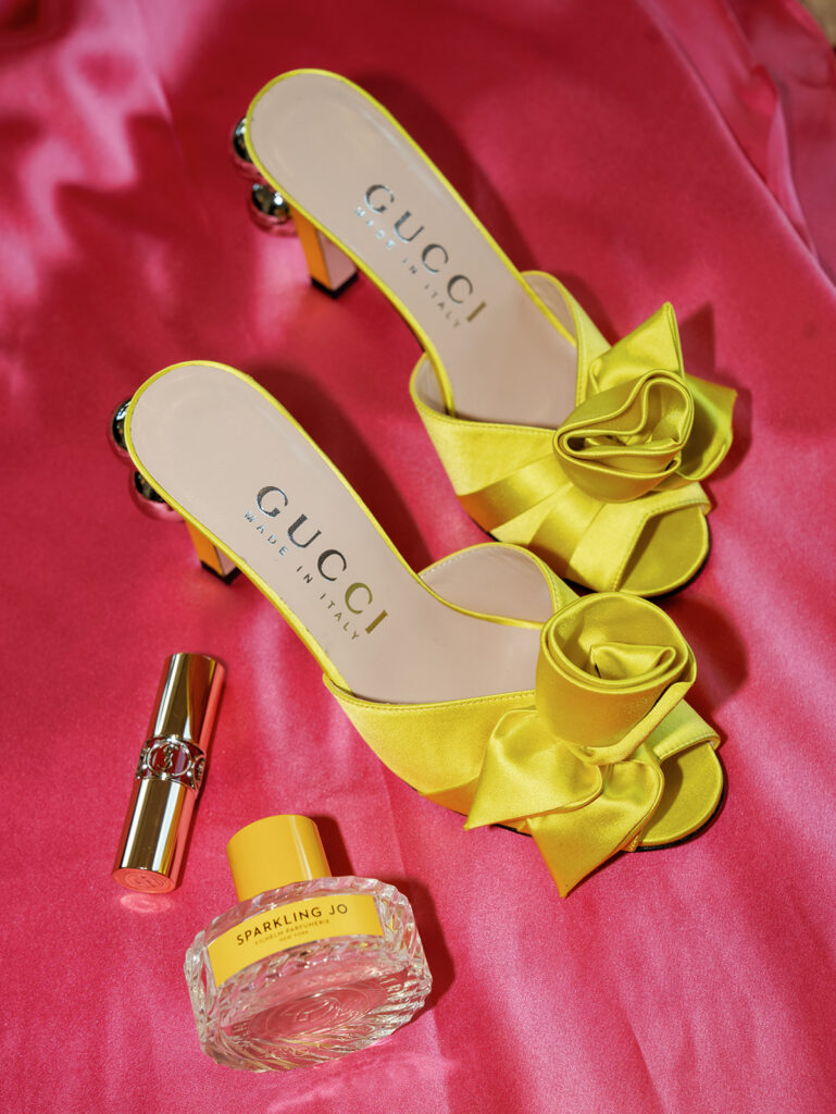 A flat lay of the brides shoes, perfume, and lipstick against a hot pink backdrop.