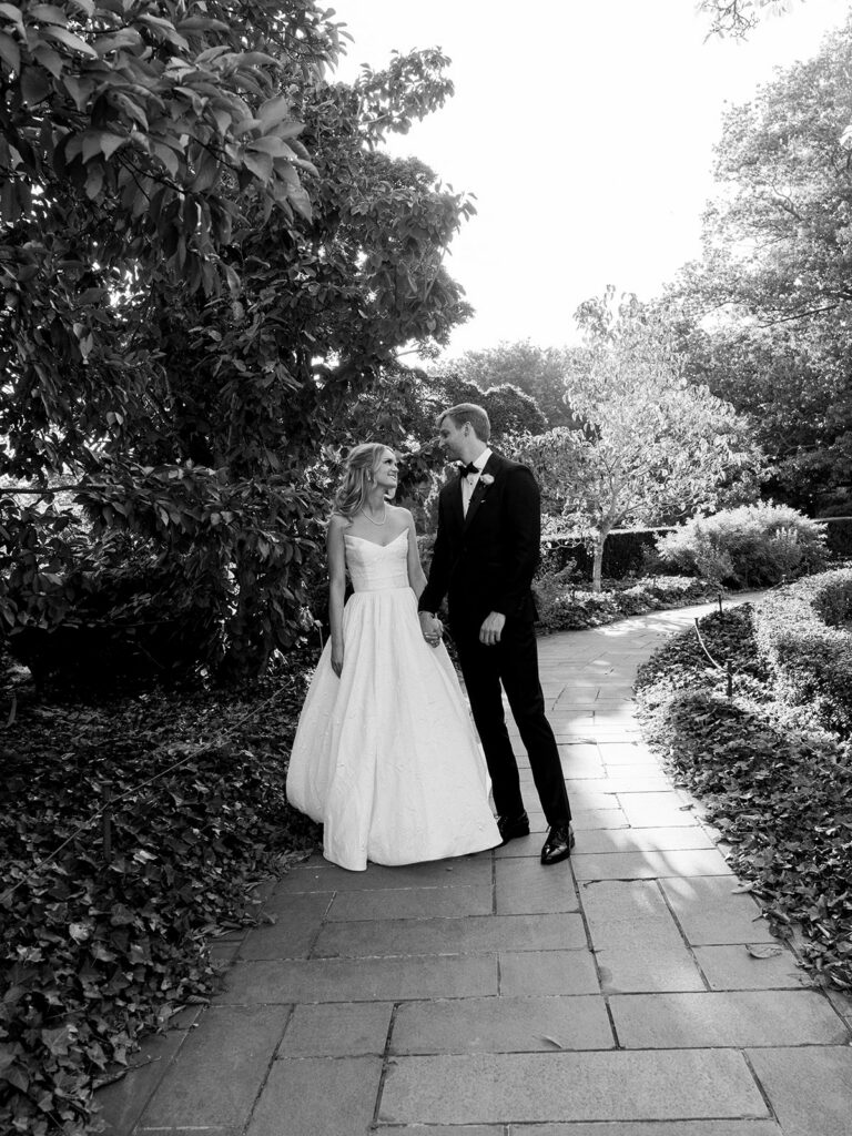 Bride and groom walking down a path together looking at each other holding hands.