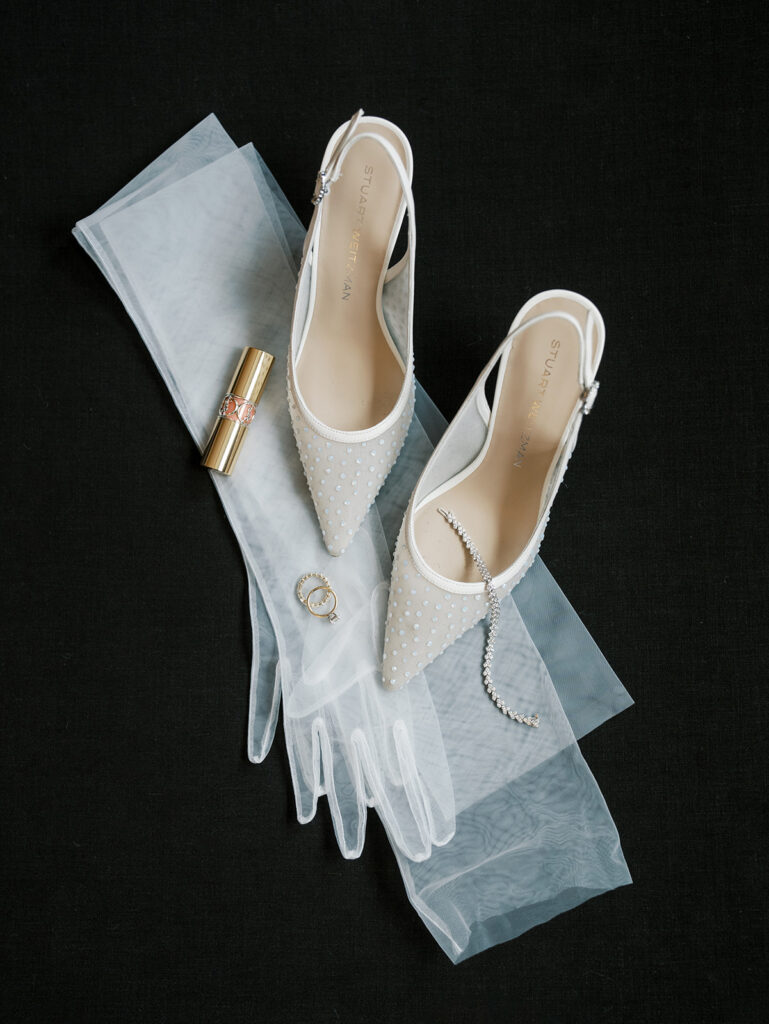 Flat lay of wedding shoes, gloves, rings and lip stick on a black background.