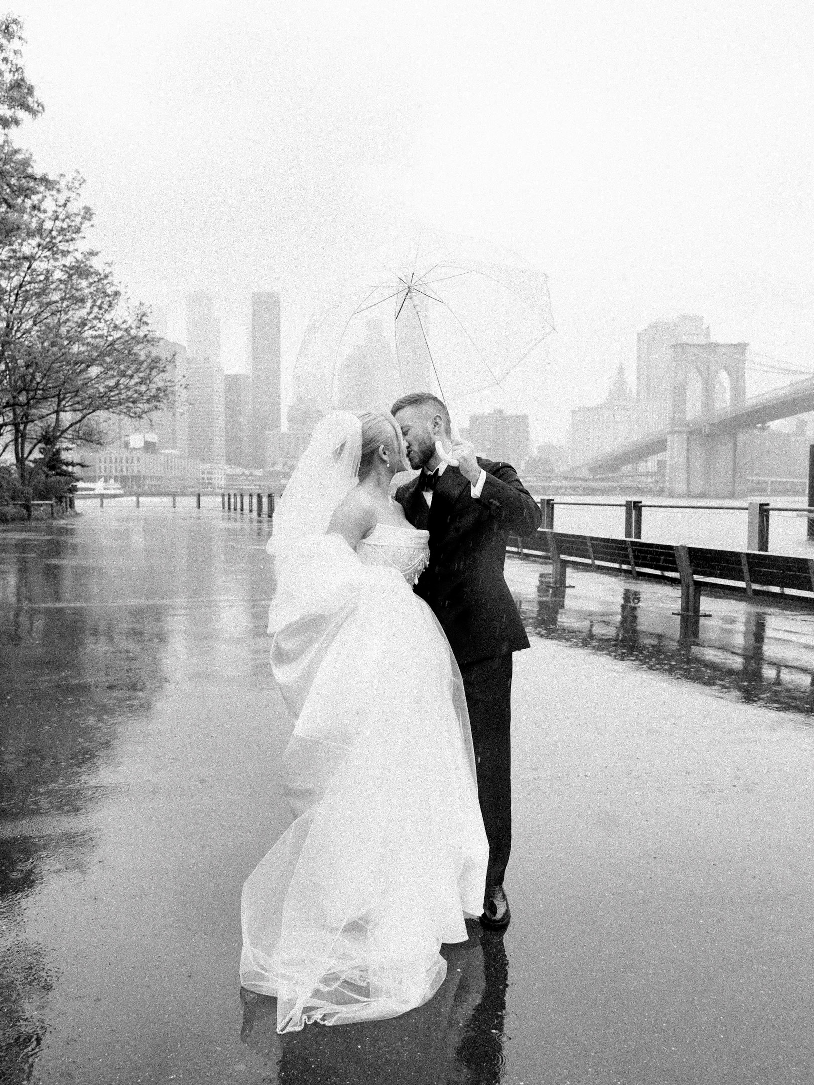 Bride and groom kissing at their rainy River Cafe wedding Brooklyn under an umbrella with the bridge view in the background.