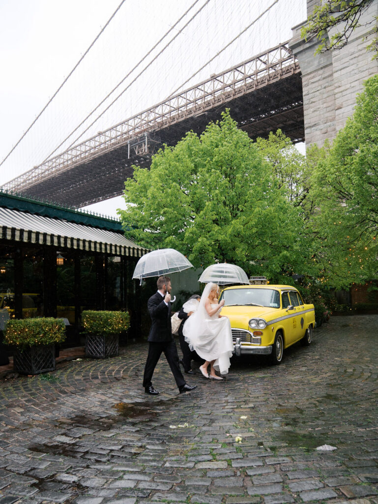 Bride and groom walking out of the river cafe in the rain to the taxi with the brooklyn bridge in the background.