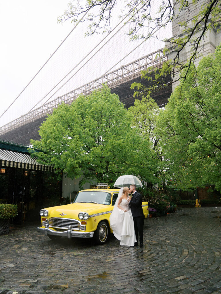 Wide image of the bride and groom against the driver's side of the yellow taxi in front of the river cafe with the brooklyn bridge in the background.