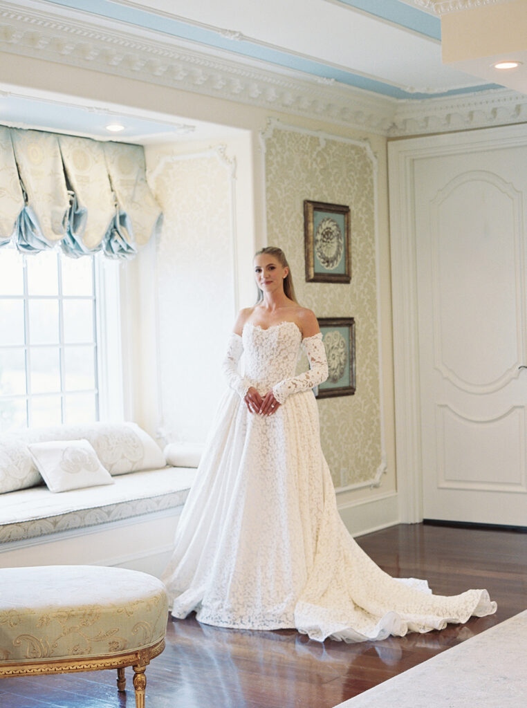 A bride standing by the window for a portrait holding her hands at her waist looking at the camera.
