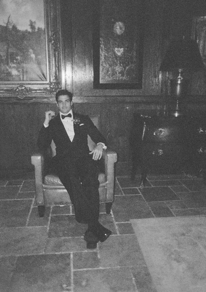 Groom sitting relaxed in a chair with one hand up near his chin looking at the camera in the library.