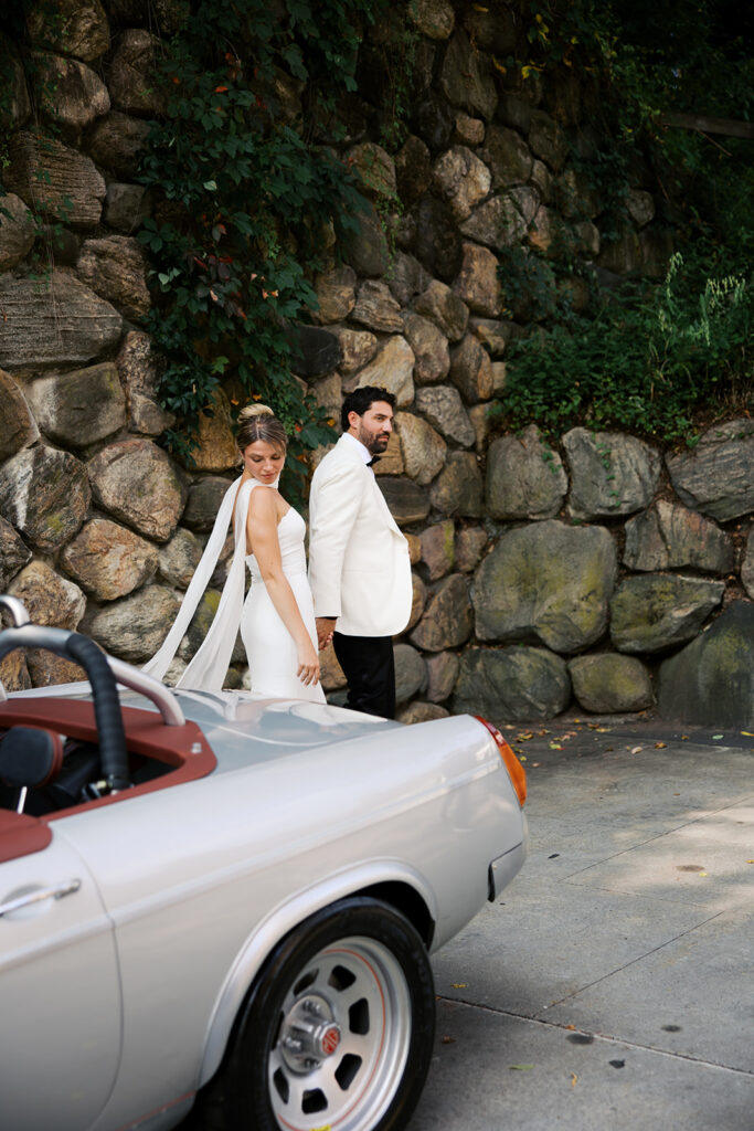 A candid walking photo of the bride and groom walking around the car with the car in the foreground.